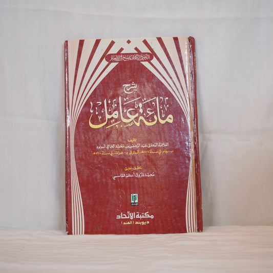 Sharh Mi'at 'Aamil" - A detailed Arabic grammar book with a red hardcover. The title and author's name are written in golden Arabic script. The book is placed against a plain white fabric background. Publisher: Maktabat Al-Ittihad, Mumbai.